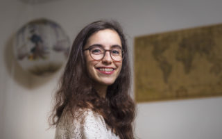 A portrait of a young woman with thick framed glasses, brown long hair, and braces.