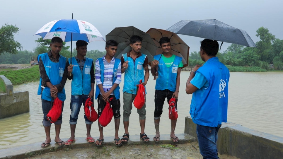 Five young Rohingya men wearing blue vests and carrying umbrellas stand listening to an older man who is carrying an umbrella at the edge of a stream in the rain