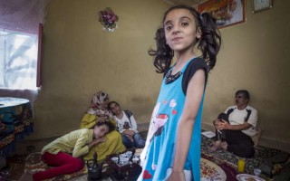 Nora (eight years old, in blue dress) is the youngest of the three daughters of Husam and Nemah. In the background, sitting on a mattress, left to right: daughter Kholod (11 years old), mother Nemah and Asmaa (14 years old). Father Husam is sitting on the right.