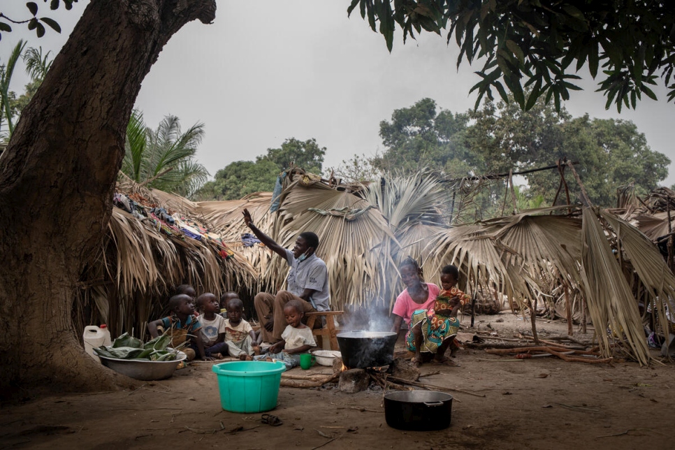 Paul sits with his family as Pascaline prepares food over a fire outside their shelter.