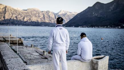 Two men sit with their backs to the camera. They are looking at a lake with mountains in the background