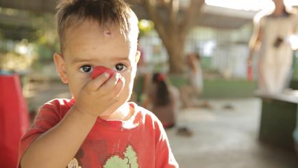 A child looks at the camera, has one hand in front of their face