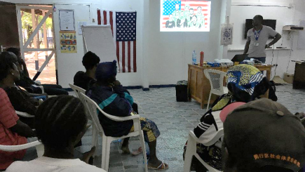 People seating on chairs in a room, listening to someone giving a presentation.