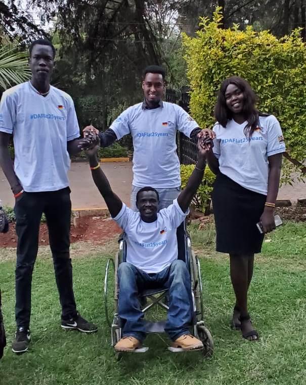 A man in a wheelchair is in front of three people. They smile at the camera.