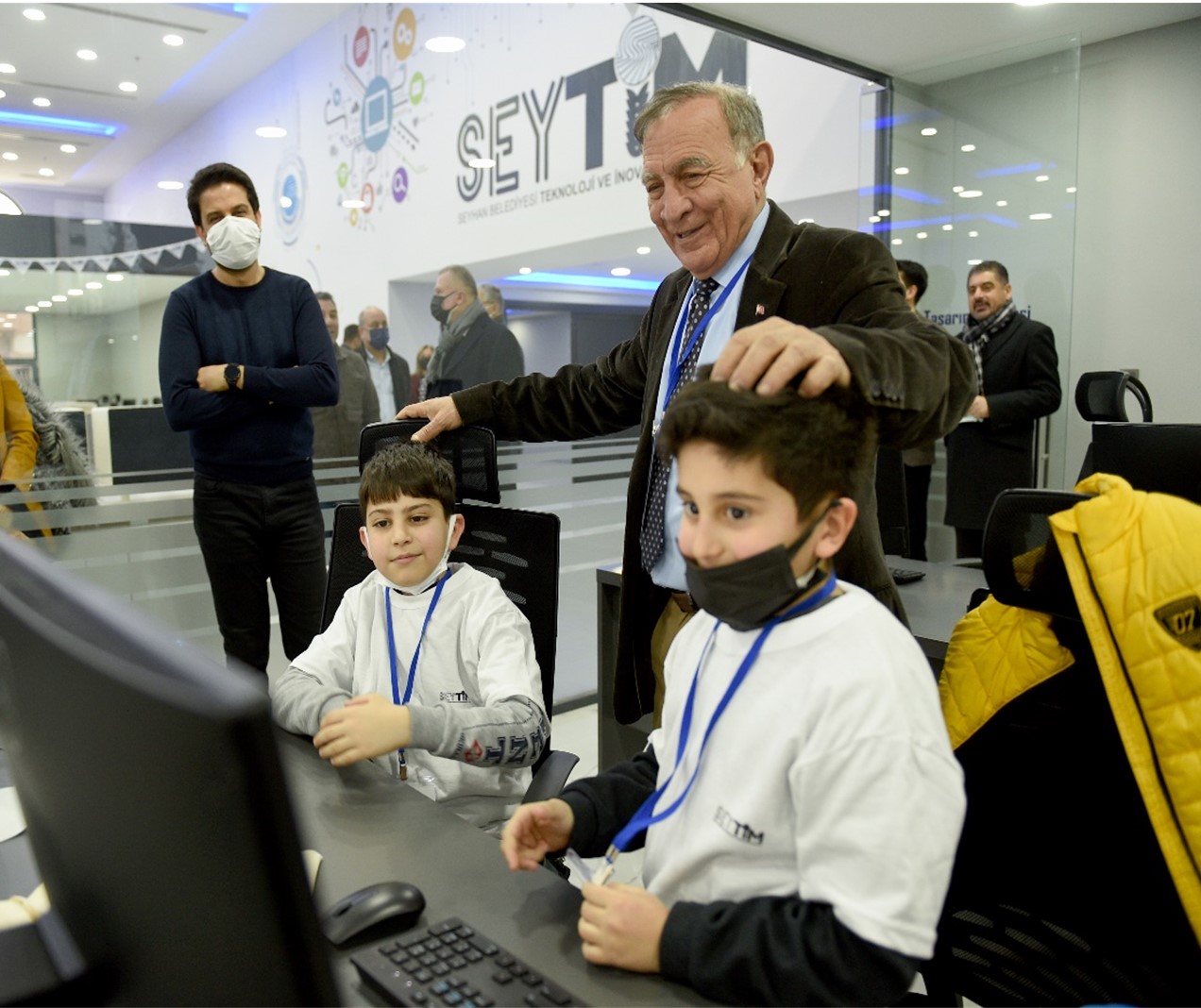 Two children with face masks sit in front of computers and are surrounded by adults