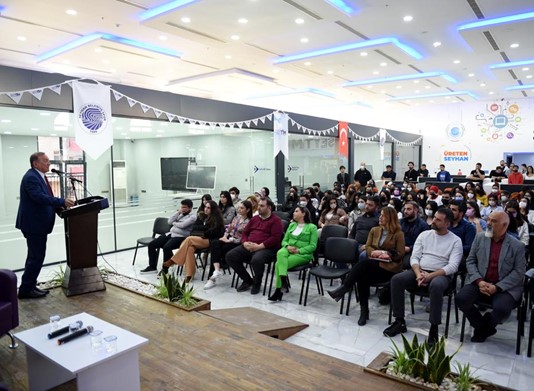 An audience listens to a man speaking at a podium in the SEYTIM building