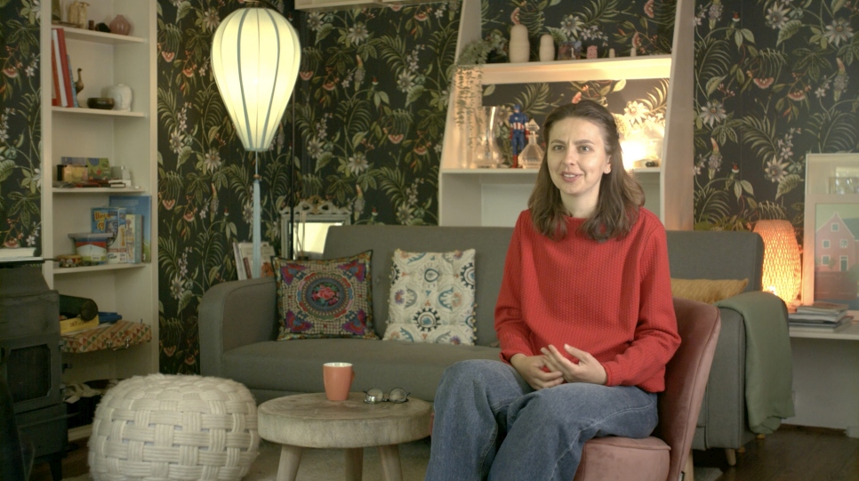 A woman with long brown hair and a red shirt sits in a living room with floral wallpaper.