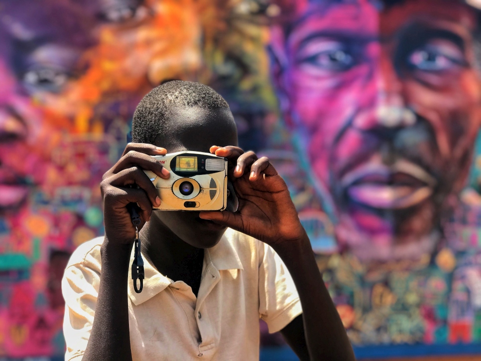 It's 8am at Alaba Primary School in Bidibidi settlement and, after reading out the news bulletin at morning assembly, young South Sudanese refugee Denis – who wants to be a journalist – takes a picture with the camera given to him by his uncle. ; Denis is the school journalist. He has a team of three students who he has trained to gather stories. They live in different blocks within zone two, which helps in preparing comprehensive news coverage from across the settlement. After a ceremony to mark World Refugee Day, Denis delivered a bulletin to the pupils and teachers at Alaba school. The mural behind him was painted by South Sudanese refugees trained by the Artolution NGO. It signifies the three generations of South Sudan affected by war – children, parents and grandparents. Uganda is home to over 1.3 million refugees, including some 865,000 from neighbouring South Sudan.