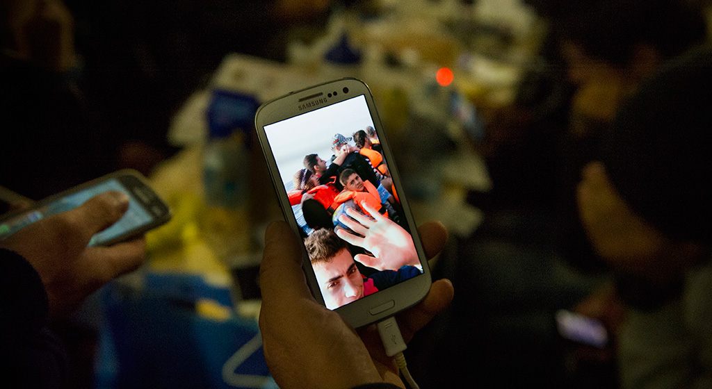 Man showing a video he made of his family crossing from Turkey to Greece in a small boat. He is charging his phone along with many others in a tent provided by Greenpeace. It also has free wifi, so becomes an important role in allowing people to contact relatives and friends, many of hwom have become separated along the route.