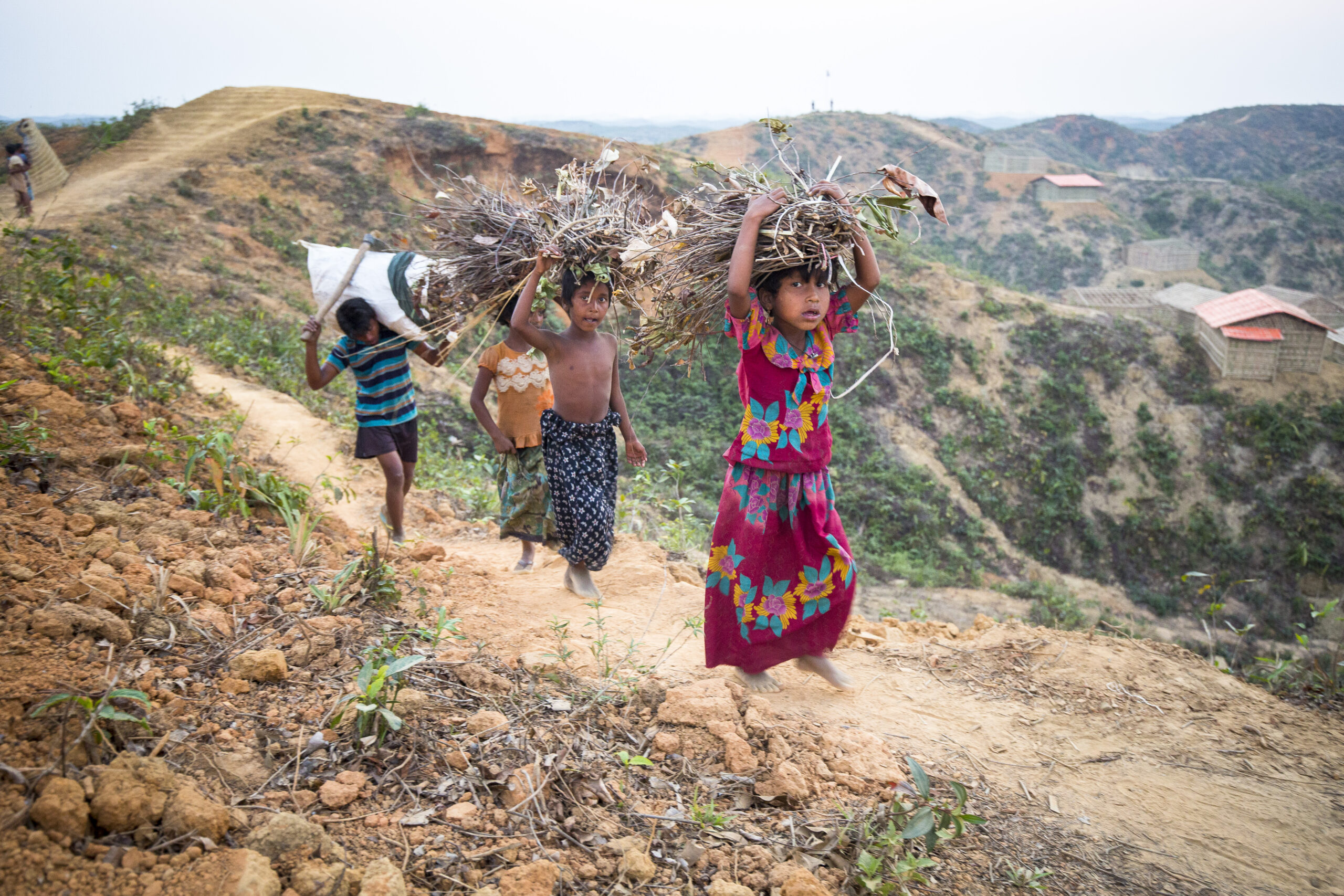 Habiba Begum, 7, in colorful floral dress, Nur Maiza, 8, wearing no shirt, and Maimuna Bibi, 8, in orange top with green skirt collecting firewood on the edge of the forest in Kutupalong Refugee Camp. © UNHCR/Roger Arnold