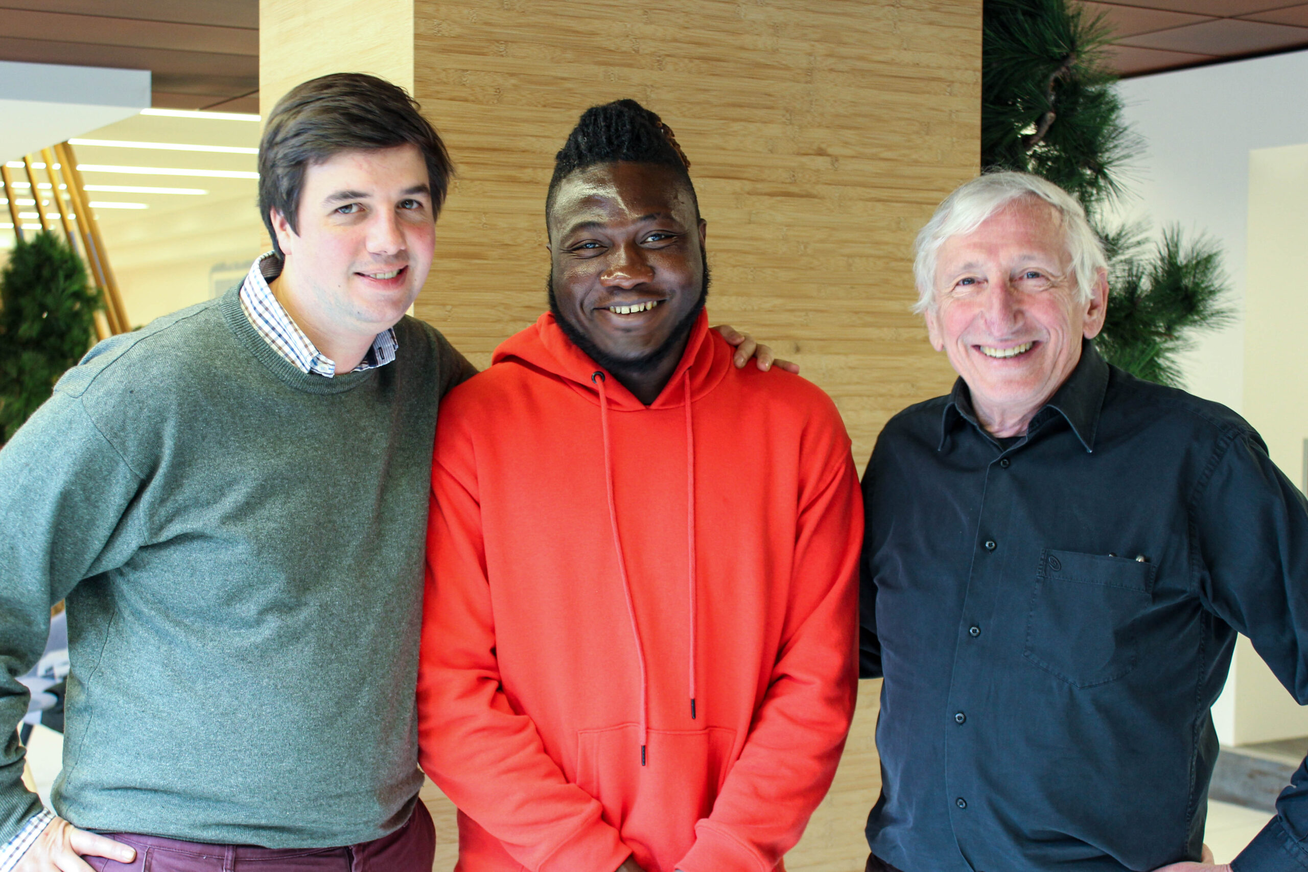 Destin, jeune réfugié camerounais, est debout aux côtés de ses deux parrains, Pierre et Quentin. Ils sourient tous les trois.