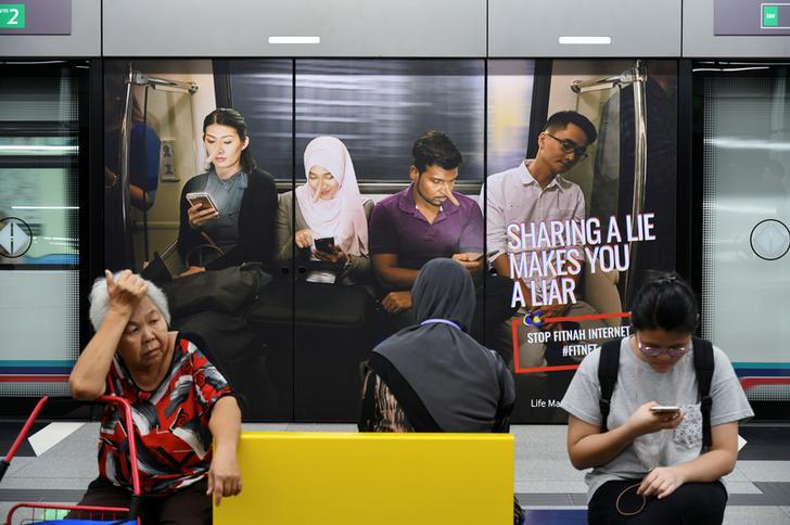 Commuters sit in front of an advertisement discouraging the dissemination of fake news, at a train station in Kuala Lumpur, Malaysia on March 28, 2018.